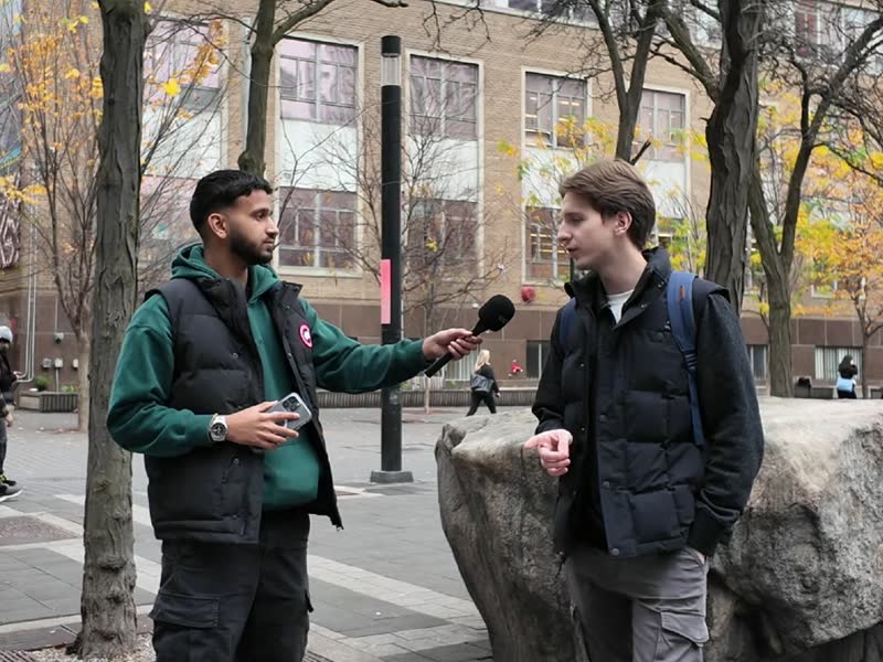 A street interview in progress: interviewer holding a microphone to a passer-by outdoors.